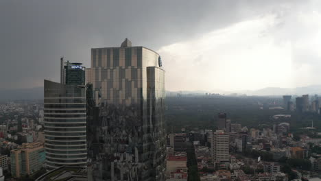 Drone-camera-rising-and-flying-towards-tall-office-glass-modern-building.-Dramatic-cloudy-sky-before-heavy-rain.-Mexico-city,-Mexico.