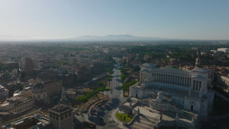 Backwards-fly-above-Piazza-Venezia-square-min-historic-city-centre.-Aerial-view-of-famous-tourist-sights,-Vittoriano-and-Colosseum-in-distance.-Rome,-Italy