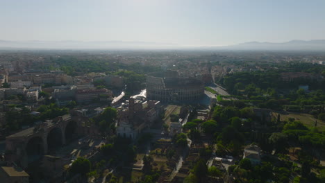 Fly-above-historic-borough-of-metropolis,-remains-of-ancient-temples-and-buildings.-Famous-Colosseum-amphitheatre-against-sun.-Rome,-Italy