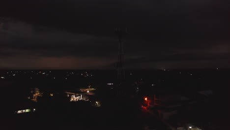 Spooky-scene-of-telecommunication-mast-with-antennas-against-dark-sky-with-flashing-lightning-during-storm.-Night-town-in-background.-Valladolid,-Mexico