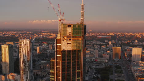 Fly-around-new-modern-skyscraper-construction-site-with-cranes-high-above-ground.-Evening-golden-hour.-Warsaw,-Poland