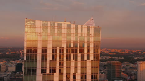 Slide-and-pan-footage-of-modern-building-with-glossy-glass-facade-reflecting-sunset-sky.-Revealing-spire-on-top-of-PKIN-building.-Warsaw,-Poland