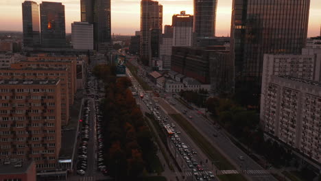 Fly-above-wide-busy-street.-Tilt-up-reveal-of-tall-downtown-skyscrapers-against-colourful-sunset-sky.-Warsaw,-Poland