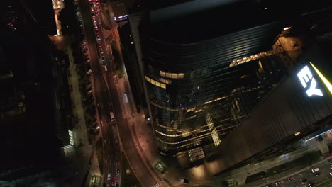 High-angle-rising-footage-of-tall-modern-skyscraper-and-traffic-on-surrounding-streets.-Night-scene.-Warsaw,-Poland