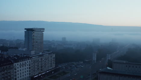 Slide-and-pan-footage-of-road-lined-with-apartment-buildings-and-public-park.-Blue-toned-misty-morning-in-city.-Warsaw,-Poland