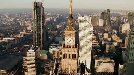 Aerial-descending-footage-of-spire-with-tower-clock-on-top-of-high-rise-PKIN-building.-Modern-tall-buildings-in-background.-Warsaw,-Poland