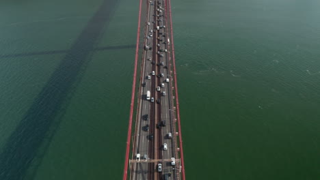 Aerial-overhead-view-following-a-busy-traffic-driving-across-the-sea-on-large-red-suspension-Ponte-25-de-Abril-bridge-in-Lisbon,-Portugal
