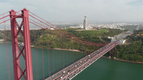 Aerial-drone-view-of-southern-end-of-25th-of-April-Bridge-connecting-Lisbon-and-Almada.-Heavy-traffic-on-multilane-road-in-rush-hour.-Big-statue-of-Jesus-on-pedestal.-Lisbon,-capital-of-Portugal.