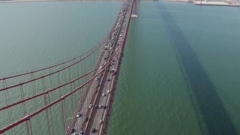 Aerial-tilt-reveal-of-busy-car-traffic-on-multi-lane-road-crossing-red-Ponte-25-de-Abril-bridge-towards-urban-city-center-of-Lisbon,-Portugal