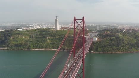 Aerial-view-of-big-red-cable-stayed-25th-of-April-Bridge-over-Tagus-river.-Multilane-road-with-heavy-traffic-from-drone.-Statue-of-Jesus-on-tall-pedestal.-Lisbon,-capital-of-Portugal.