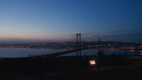 Gradually-revealing-aerial-panorama-of-cable-stayed-bridge-after-sunset.-Night-bridge-silhouette-from-drone.-Lisbon,-capital-of-Portugal.