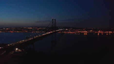 Aerial-night-dolly-out-view-of-Ponte-25-de-Abril-red-bridge-with-car-traffic-and-Lisbon-city-center-with-lights-in-the-background