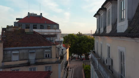 Drone-camera-flying-through-narrow-street-and-revealing-panoramic-view-on-25th-of-April-Bridge-over-Tegus-river.-Evening-elevated-footage-in-sunset-time.-Lisbon,-capital-of-Portugal.
