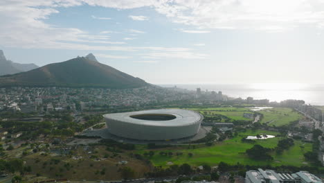 Slide-and-pan-shot-of-futuristic-structure.-Modern-football-stadium-surrounded-by-golf-course.-Buildings-in-vacation-resort-on-ocean-coast.-Cape-Town,-South-Africa