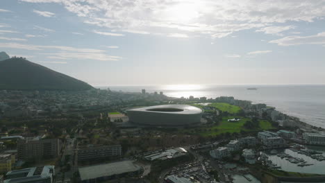 Backwards-fly-above-sea-coast-in-city-at-sunset.-Modern-structure-of-Green-Point-Stadium-and-surrounding-development.-Cape-Town,-South-Africa