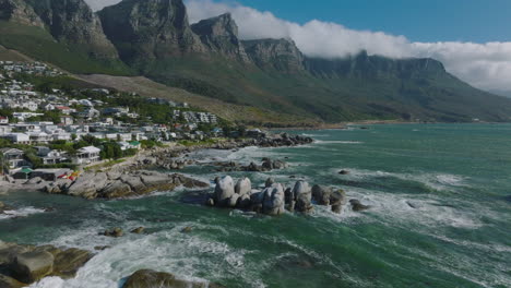 Fly-above-coast.-Rough-sea-with-waves-crashing-to-coastal-rocks.-Residential-suburb-under-high-mountain-ridge.-Cape-Town,-South-Africa