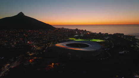 Modern-football-stadium-and-surrounding-town-development-after-sunset.-Urban-borough-at-seaside-at-dusk.-Cape-Town,-South-Africa