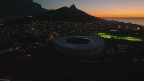 Slide-and-pan-aerial-footage-of-city-with-modern-sports-arena.-Panoramic-view-of-town-at-seaside-and-mountains-against-twilight-sky.-Cape-Town,-South-Africa