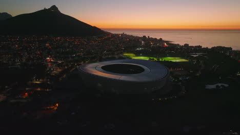 Fly-above-city-in-evening.-Modern-football-arena-and-streetlights-in-surrounding-urban-borough.-Colourful-sunset-sky.-Cape-Town,-South-Africa