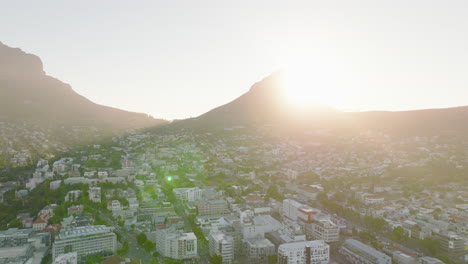 Forwards-descending-fly-above-buildings-in-urban-borough.-View-against-bright-sun,-gradually-hiding-of-glow-behind-mountain-peak.-Cape-Town,-South-Africa