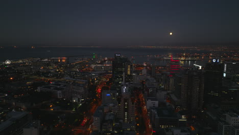 Forwards-fly-above-row-of-high-rise-buildings-in-city-centre.-Sea-bay-with-big-seaport-on-coast-at-night.-Cape-Town,-South-Africa