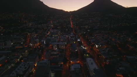 Night-angle-view-of-illuminated-streets-in-night-town.-Tilt-up-reveal-of-mountain-silhouettes-against-twilight-sky.-Cape-Town,-South-Africa