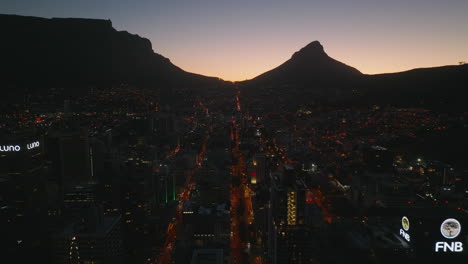 Fly-above-night-city.-Silhouette-of-mountains-against-picturesque-twilight-sky.-Backwards-revealing-of-high-rise-downtown-office-buildings.-Cape-Town,-South-Africa