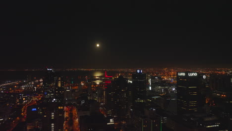 Aerial-descending-footage-of-night-in-central-part-of-city.-Tall-business-buildings-and-water-surface-in-sea-bay-reflecting-moonlight.-Cape-Town,-South-Africa