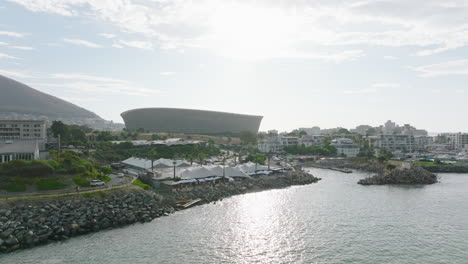 Slide-and-pan-shot-of-modern-Green-Point-football-stadium,-Fly-along-sea-coast-with-restaurant.-Cape-Town,-South-Africa