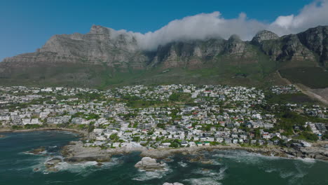 Aerial-ascending-footage-of-houses-in-Bakoven-residential-suburb.-Buildings-in-slope-rising-from-sea-coast-to-beautiful-rocky-mountain-ridge.-Cape-Town,-South-Africa