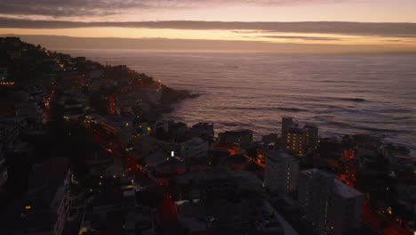 Fly-above-buildings-in-urban-neighbourhood-at-sea-coast-at-dusk.-Rippled-water-surface-and-colourful-sky.-Cape-Town,-South-Africa