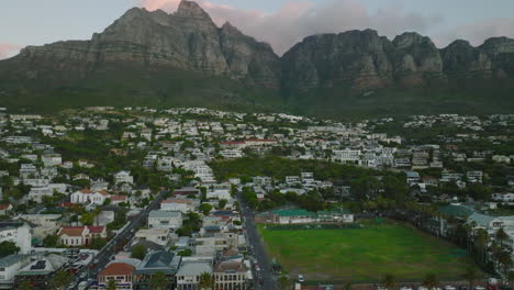 Slide-and-pan-aerial-shot-of-urban-neighbourhood.-Low-buildings-under-monumental-rocky-mountain-ridge-at-twilight.-Cape-Town,-South-Africa