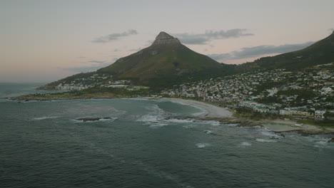 Backwards-fly-above-sea-surface-near-coast.-View-of-seaside-city-and-step-slope-Loins-Head-Mountain.-Cape-Town,-South-Africa