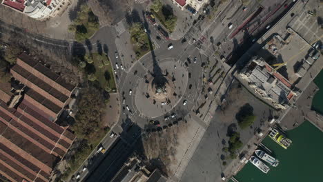 Overhead-footage-of-stream-of-vehicles-passing-on-road-around-Columbus-Monument-on-waterfront-at-port.-Barcelona,-Spain