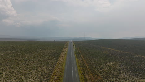 Straight-road-in-barren-landscape-with-various-shrubs.-Forwards-fly-against-truck-driving-on-road.-South-Africa