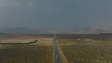 Aerial-footage-of-long-straight-road-with-low-traffic-leading-through-flat-wasteland,-mountains-in-distance.-South-Africa