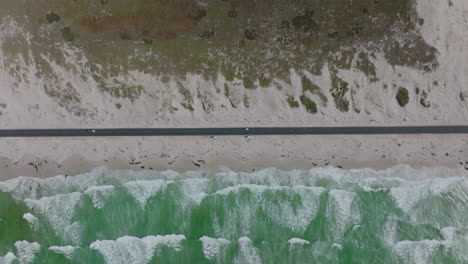 Top-down-horizontally-panning-footage-of-straight-coastal-road-passing-along-waves-washing-sand-beach.