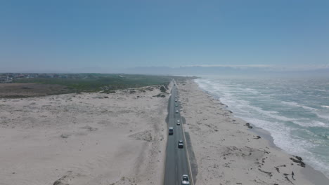 Aerial-descending-shot-of-cars-driving-on-road-along-ocean-coast.-Sea-waves-washing-sand-beach.