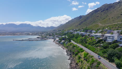 Aerial-view-of-coastal-road-leading-along-row-of-luxury-houses.-Panoramic-view-of-Gordons-Bay-with-mountains-in-background.