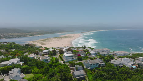 Elevated-view-of-sea-coast,-waves-rolling-to-sand-beach.-Backwards-reveal-of-luxury-residencies-surrounded-by-vegetation.-Plettenberg-Bay,-South-Africa