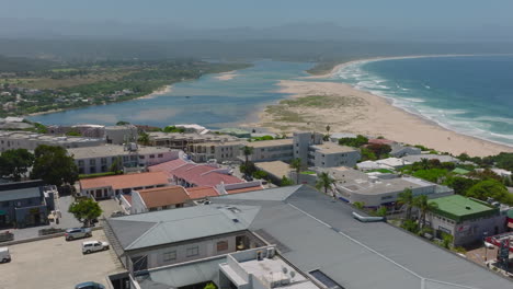 Cinematic-shot-of-buildings-around-street-in-urban-borough-high-above-sea-coast.-Water-surface-and-sandy-beach-in-background.-Plettenberg-Bay,-South-Africa