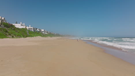 Couple-strolling-on-beach-along-rolling-waves.-Forwards-fly-along-coast,-houses-near-sea.-South-Africa