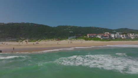 People-enjoying-sunny-day-on-sand-beach.-Backwards-reveal-of-water-surface-and-waves-rolling-to-coast.-South-Africa
