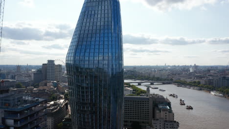 Crane-up-shot-of-One-Blackfriars-modern-apartment-building.-Glossy-glass-facade-of-skyscraper-reflecting-sky.-Aerial-view-of-Thames-river-flowing-through-city.-London,-UK