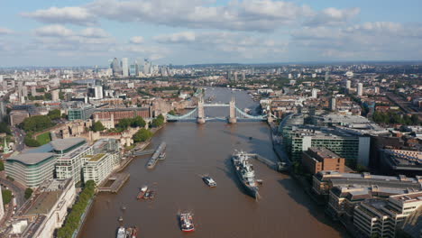 Aerial-view-of-old-famous-Tower-Bridge-across-River-Thames.-HMS-Belfast-museum-ship-moored-at-City-hall.-Modern-skyscrapers-in-Canary-Wharf-financial-hub-in-background.-London,-UK