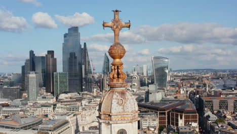 Panorama-curve-shot-footage-of-decorative-cross-on-top-of-church-tower-with-cityscape-in-background.-Group-of-tall-modern-skyscrapers-in-City-business-district.-London,-UK