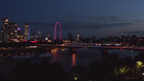 Rising-footage,-night-city-revealing-behind-trees-on-river-bank.-Color-lit-London-eye-and-bridges-over-Thames-river.-London,-UK