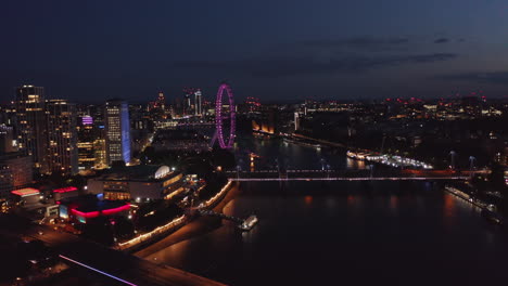 Aerial-view-of-city-after-sunset.-Night-fly-above-Thames-river,-Illuminated-bridges-and-violet-London-eye.-London,-UK