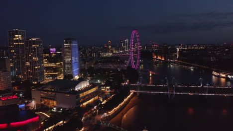 Ascending-footage-of-Thames-river-south-bank-at-night.-Royal-Festival-Hall-and-London-eye.-Aerial-view-at-colourful-city-lights.-London,-UK