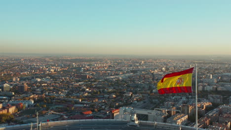Fly-around-Spanish-flag-waving-in-wind-on-rooftop-of-business-skyscraper.-Revealing-aerial-view-of-cityscape-lit-by-setting-sun.